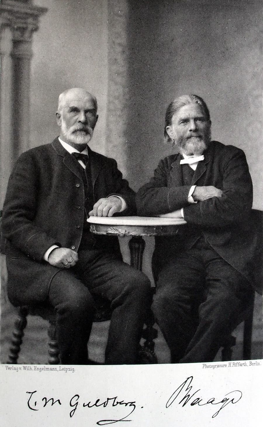 A photogravure of two elderly bearded men in dark suits seated on either side of a small round table, Guldberg on the left with a white beard and Waage on the right with his arms folded on the table. Their signatures are printed beneath.