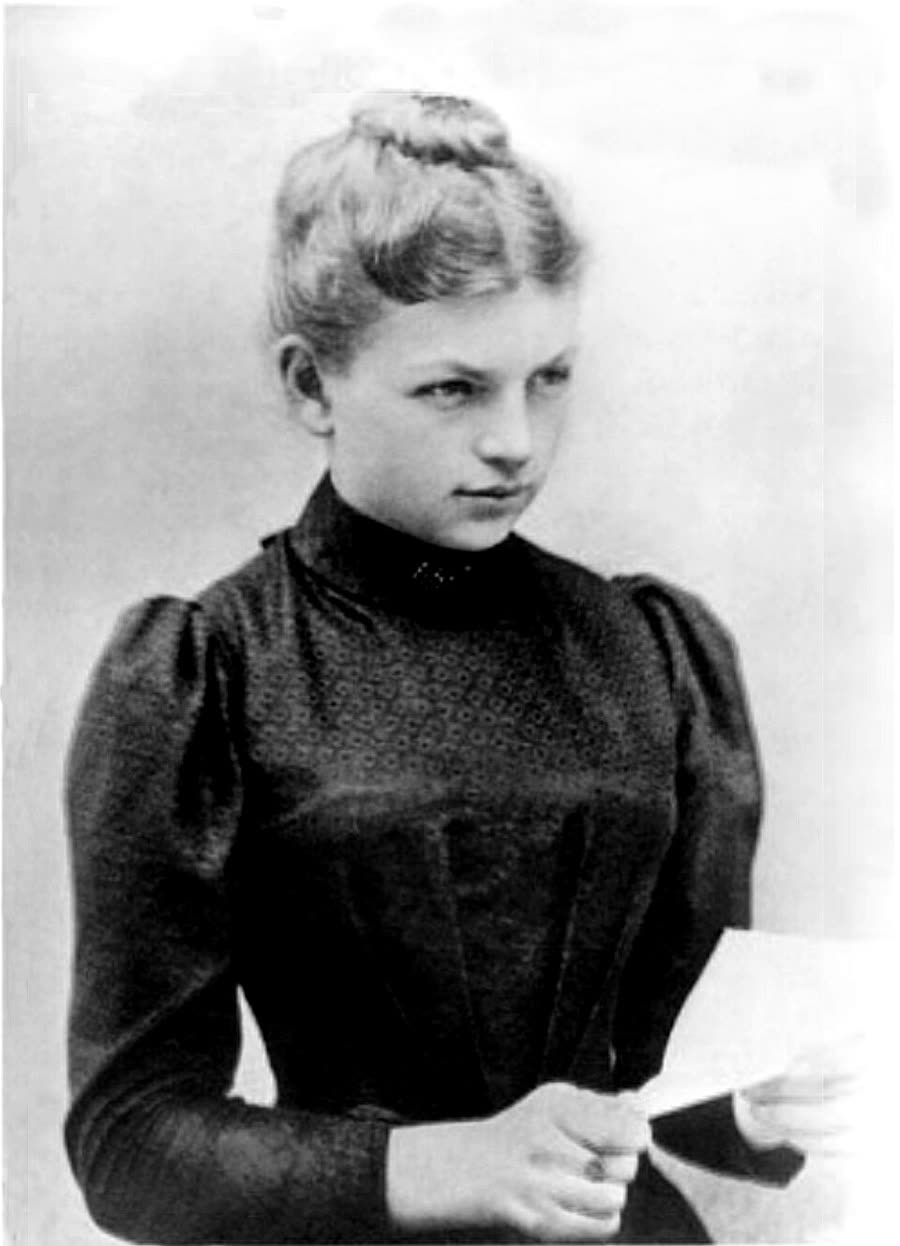 A photograph of Clara Immerwahr as a young woman, in a high-necked dark patterned dress with puffed sleeves, her fair hair pinned up, holding a sheet of paper and looking to her left.