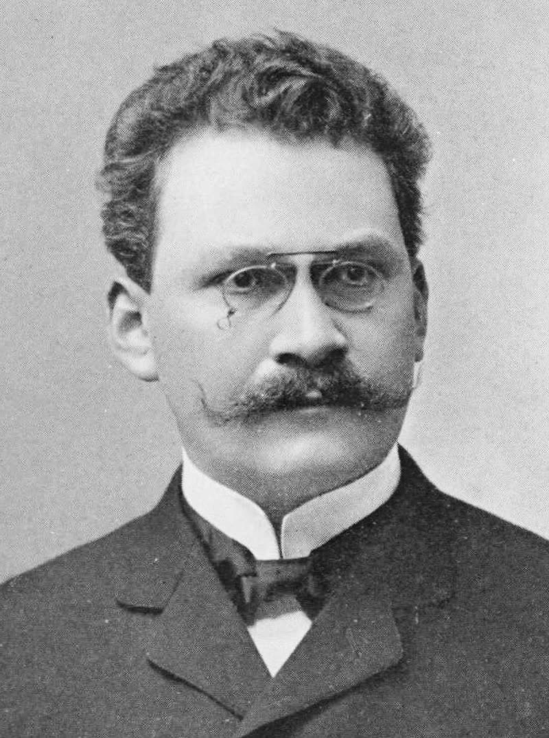 Portrait of Hermann Minkowski, seated, with a moustache and round spectacles.
